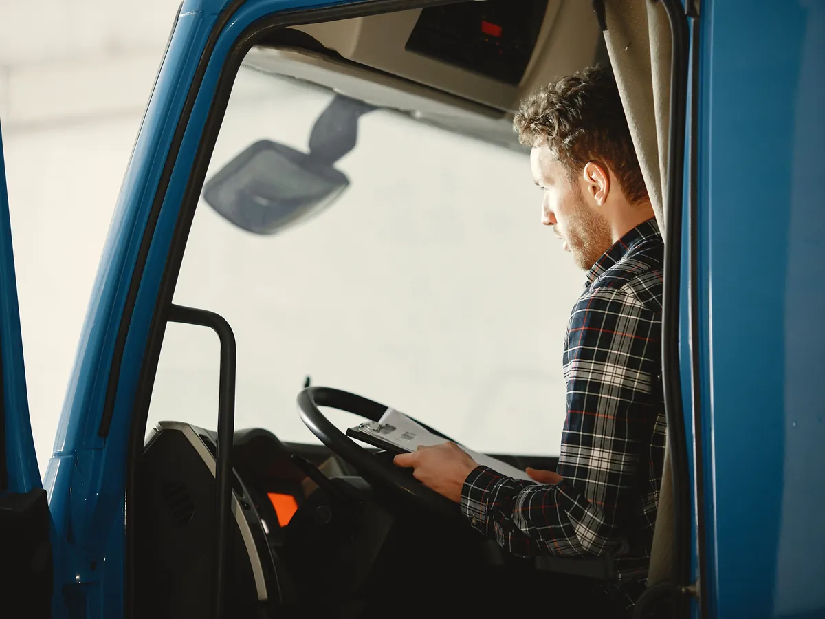 A driver in a plaid shirt checking paperwork from the cab of a blue truck.