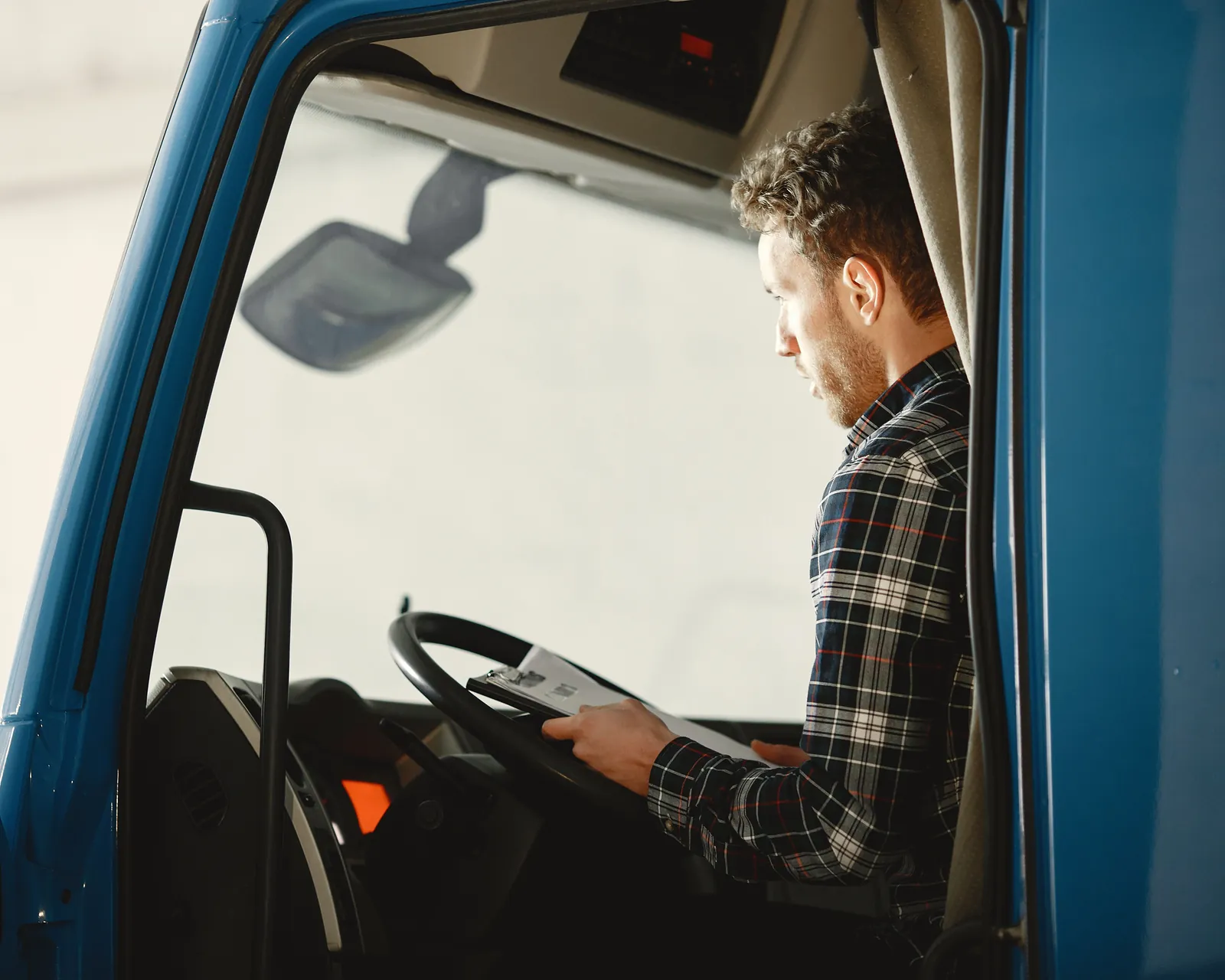 A driver in a plaid shirt checking paperwork from the cab of a blue truck.