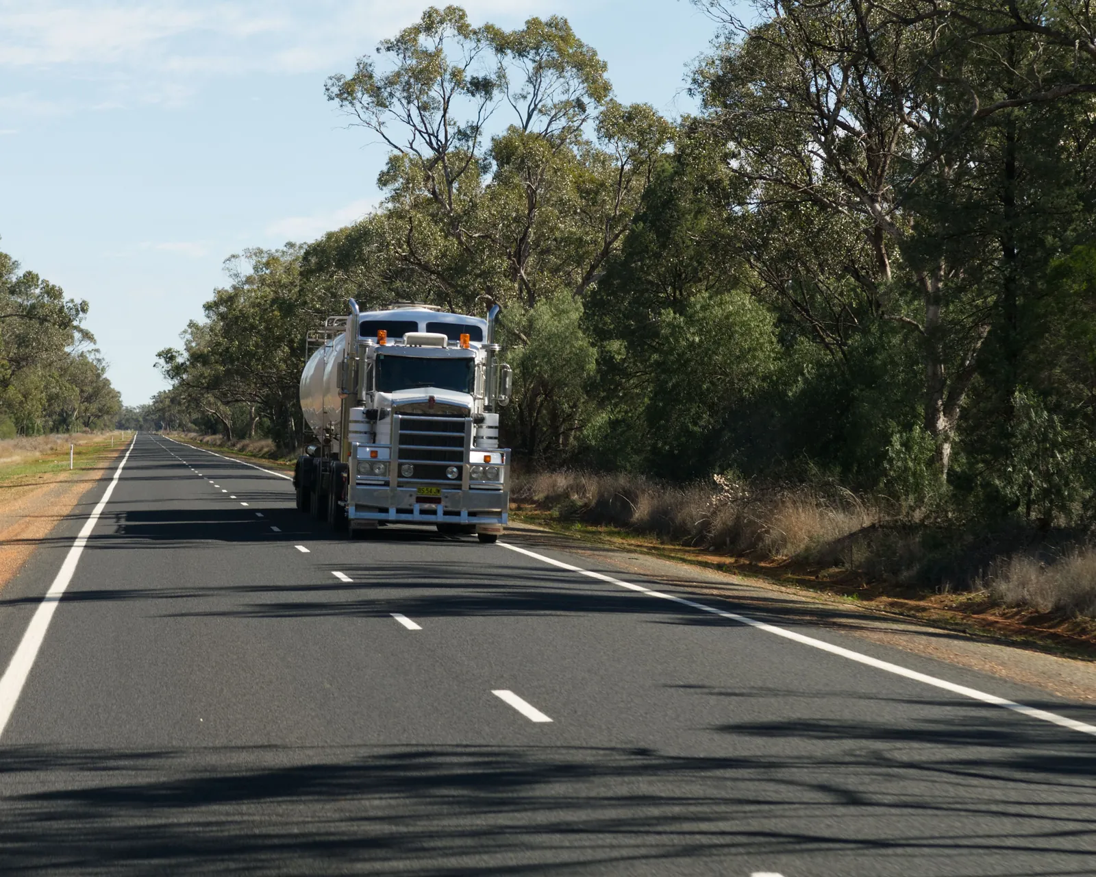 A heavy truck driving along a straight regional road lined with gum trees under a pale blue sky.