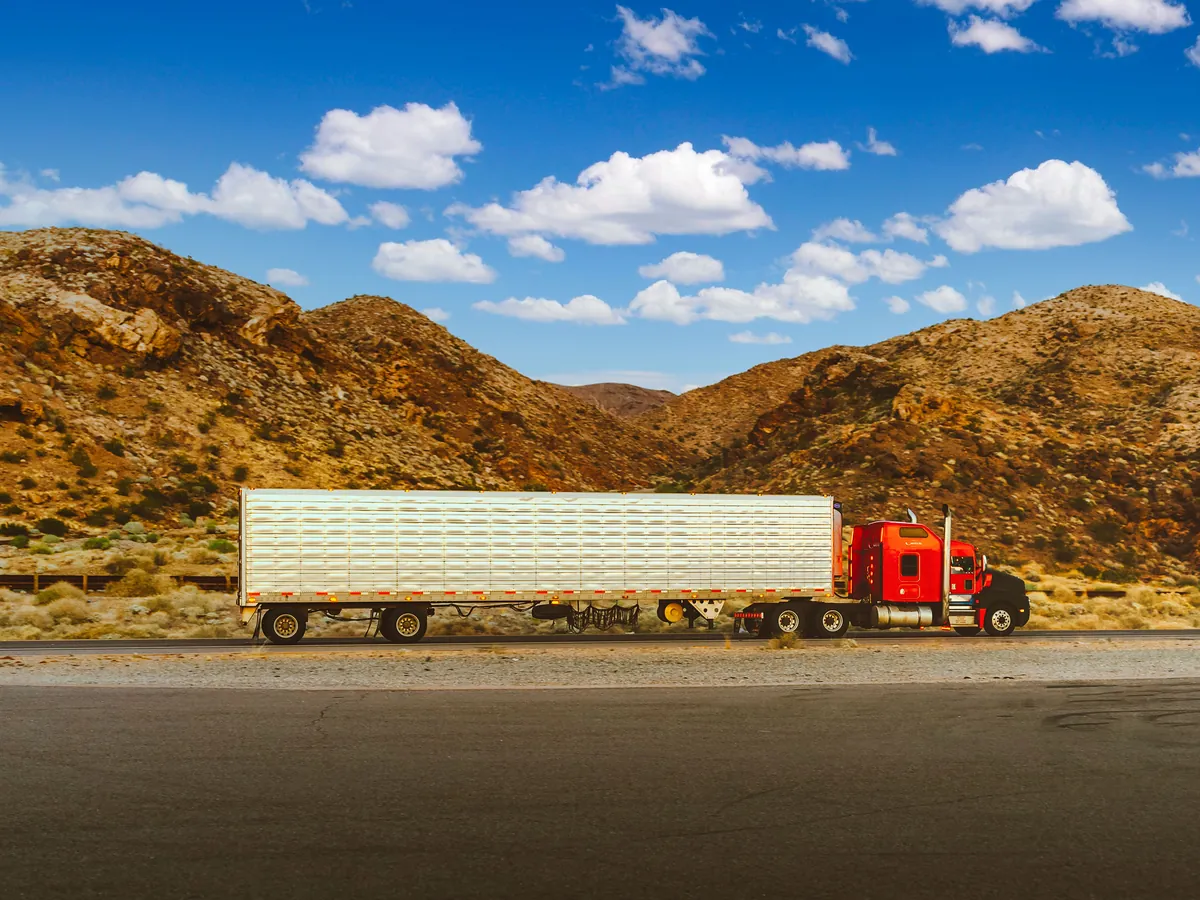 A red semi-trailer truck driving along a highway with dry hills behind it.