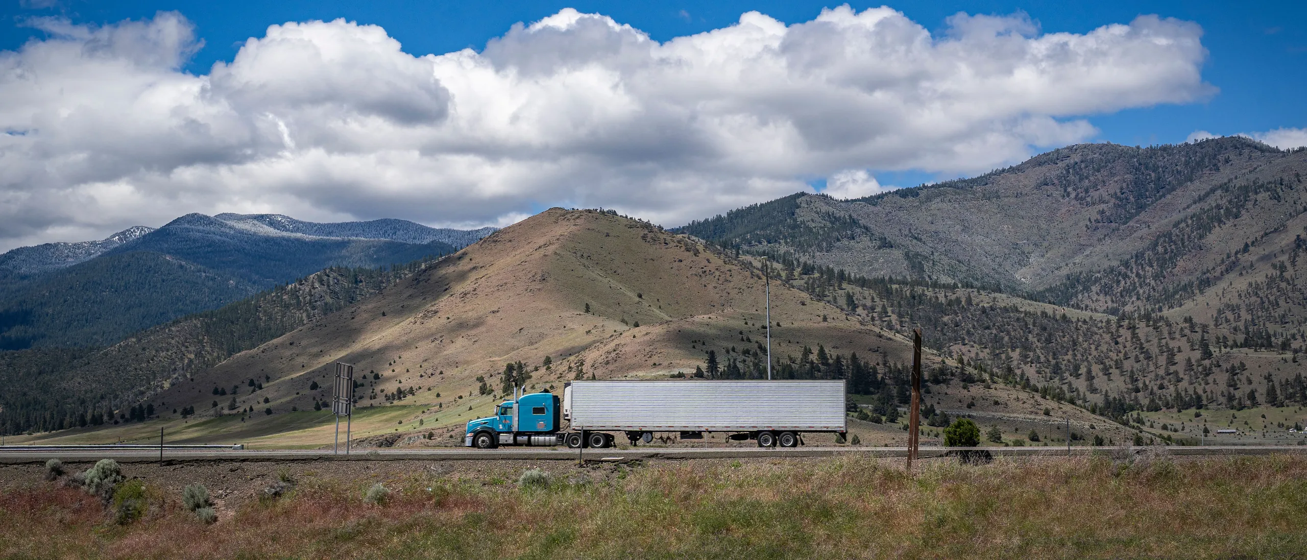 A truck travelling along a highway below dry hills and a large cloudy sky.
