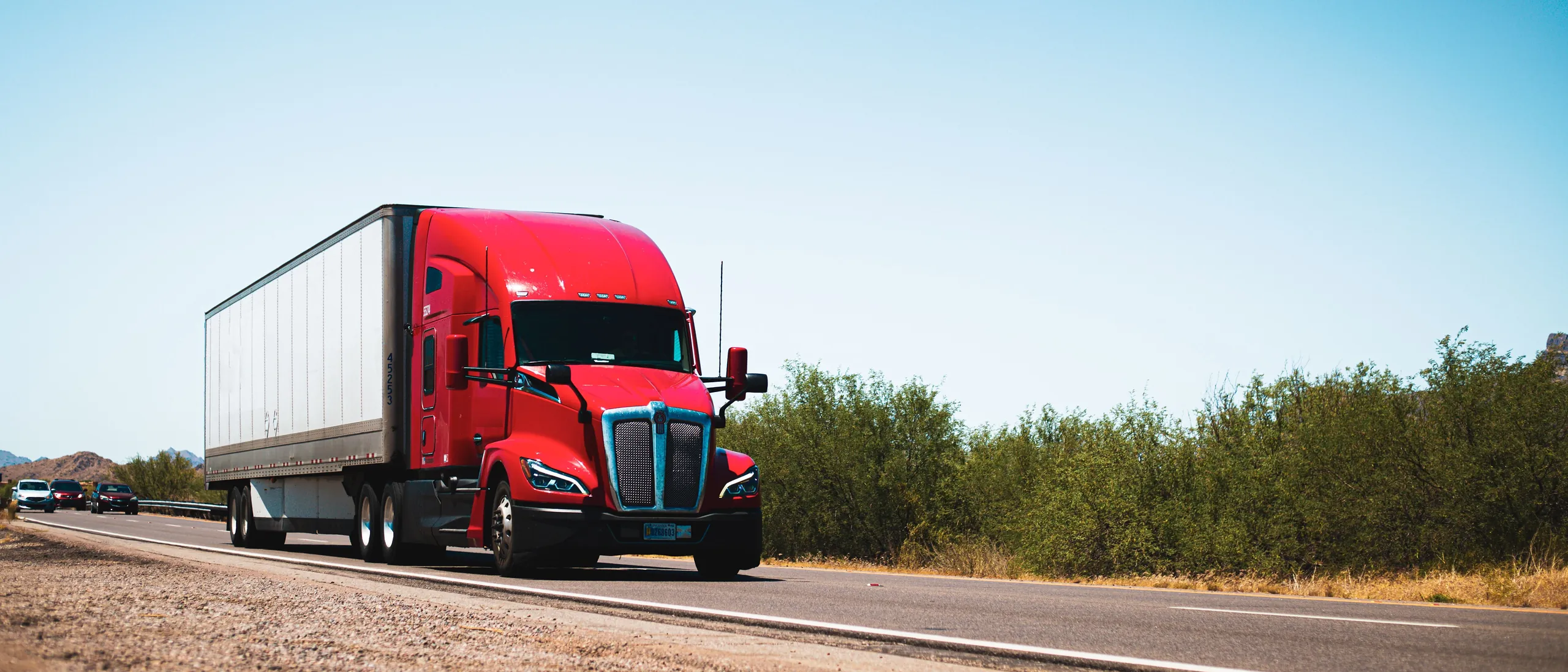 A red semi-trailer truck driving along a sunlit highway under a clear blue sky.